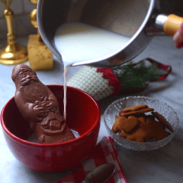 Mjölk hälls från en kastrull ner i en kopp, i koppen står en chokladtomte, i fögrunden ligger en rödrutig servett med en tesked på.