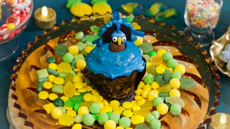 En blå påfågel i glasyr på en brun cupcake som står på gul och grön godis.