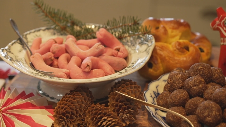 Två gamla serveringsfat i vitt och blått håller rosa marsipankorvar och den andra choklad köttbullar, faten står på en träskärbräda omgivna av lussebullar, kottar och dekorationer av papp.