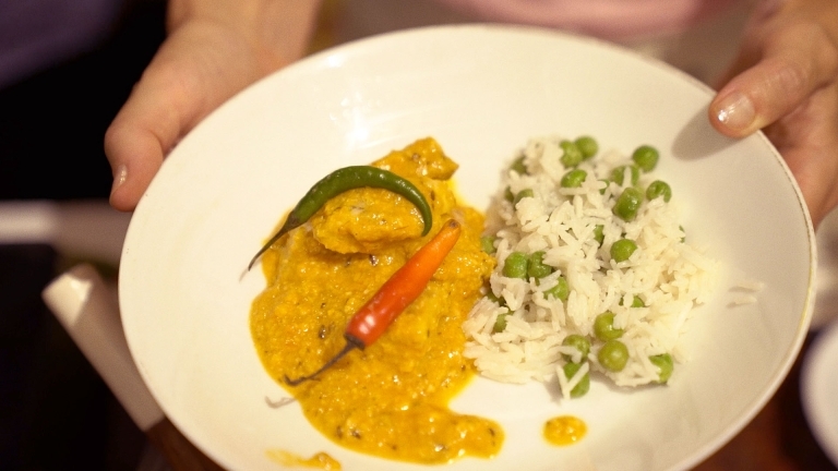 Händer håller en tallrik med ris blandat med gröna ärtor och en fisk i gul sås.