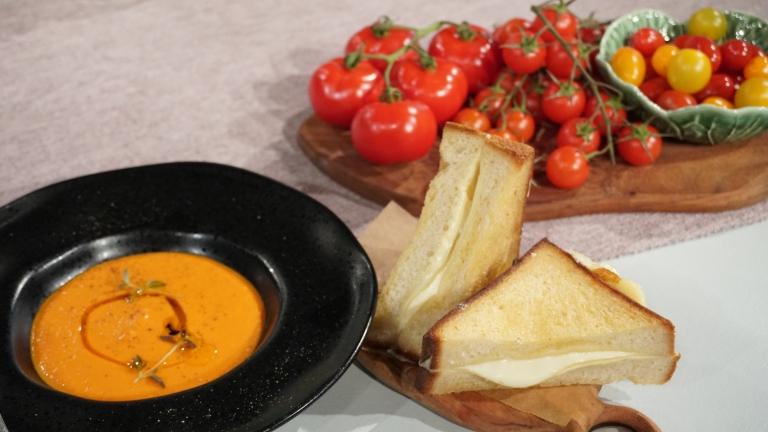 Bredvid en skål med tomatsoppa finns en skärbräda i trä, på den finns en delad ostmacka med varm ost som smälter ut mellan bröden.