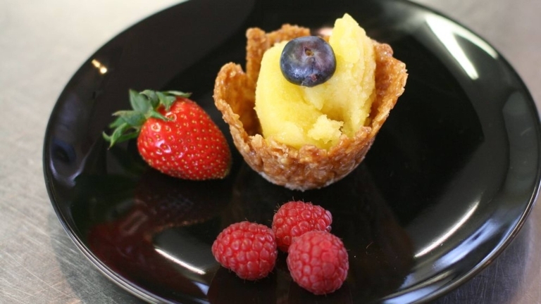 På ett svart fat ligger en jordgubbe, tre hallon och ett gyllenbrunt skal med gult sorbetinnehåll och ett blåbär på toppen. 