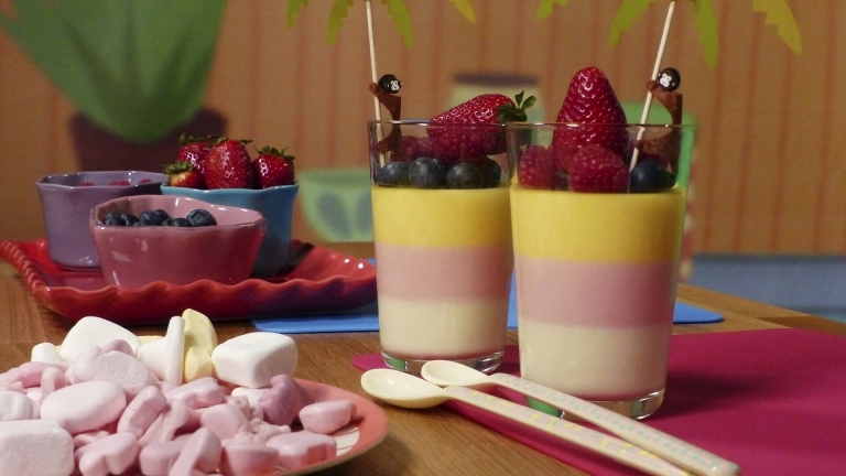 Ett fat med godis, ett fat med skålar med blåbär, jordgubbar och hallon, och två glas randig godispannacotta toppad med blåbär, jordgubbar, hallon och två små parasoll.