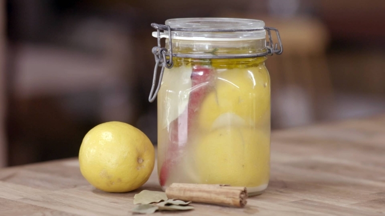 En glasburk med inlagda citroner, med en citron bredvid, kanel och lagerblad. 