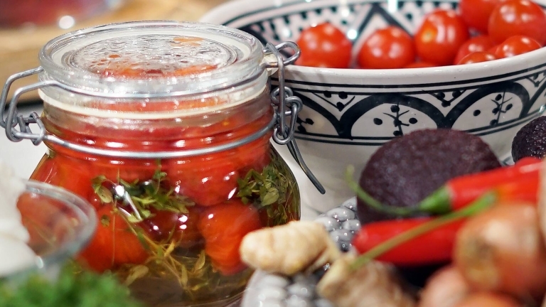 En glasburk med picklade tomater. Runt om finns andra grönsaker och ingredienser i köket. 