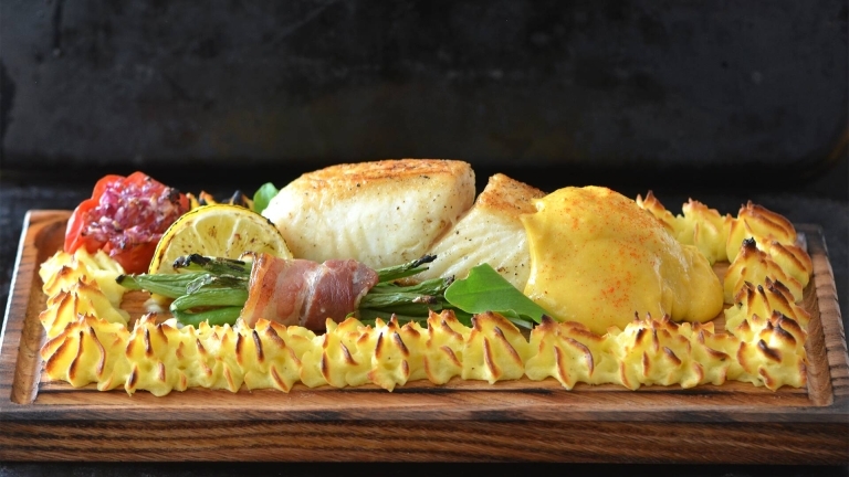 Liggande fisk, potatis mos, en bunt bönor med en baconskiva runt på en trä rektangel tallrik.