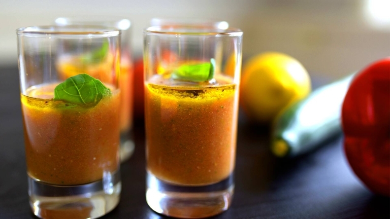 Fyra glas som är fylld av Gazpacho med en bladbasilika på varje glas och en hel gurka och citron i bakom