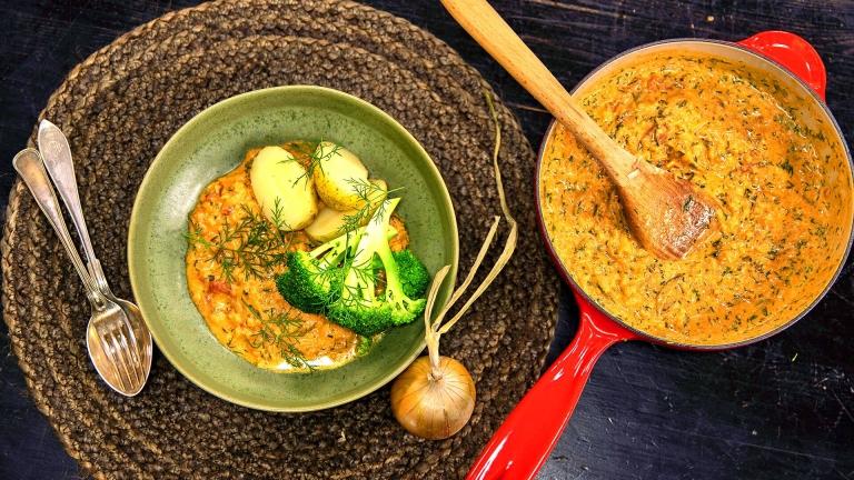 En röd stekpanna och en grön skål som är fyllda av grytan och toppning med broccoli, potatis. En sked, en gaffel  och en hel lök ligger i bredvid.