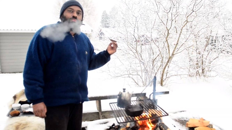 En man står utomhus och lagar mat över öppen eld. han andas rök och håller i en matbit.
